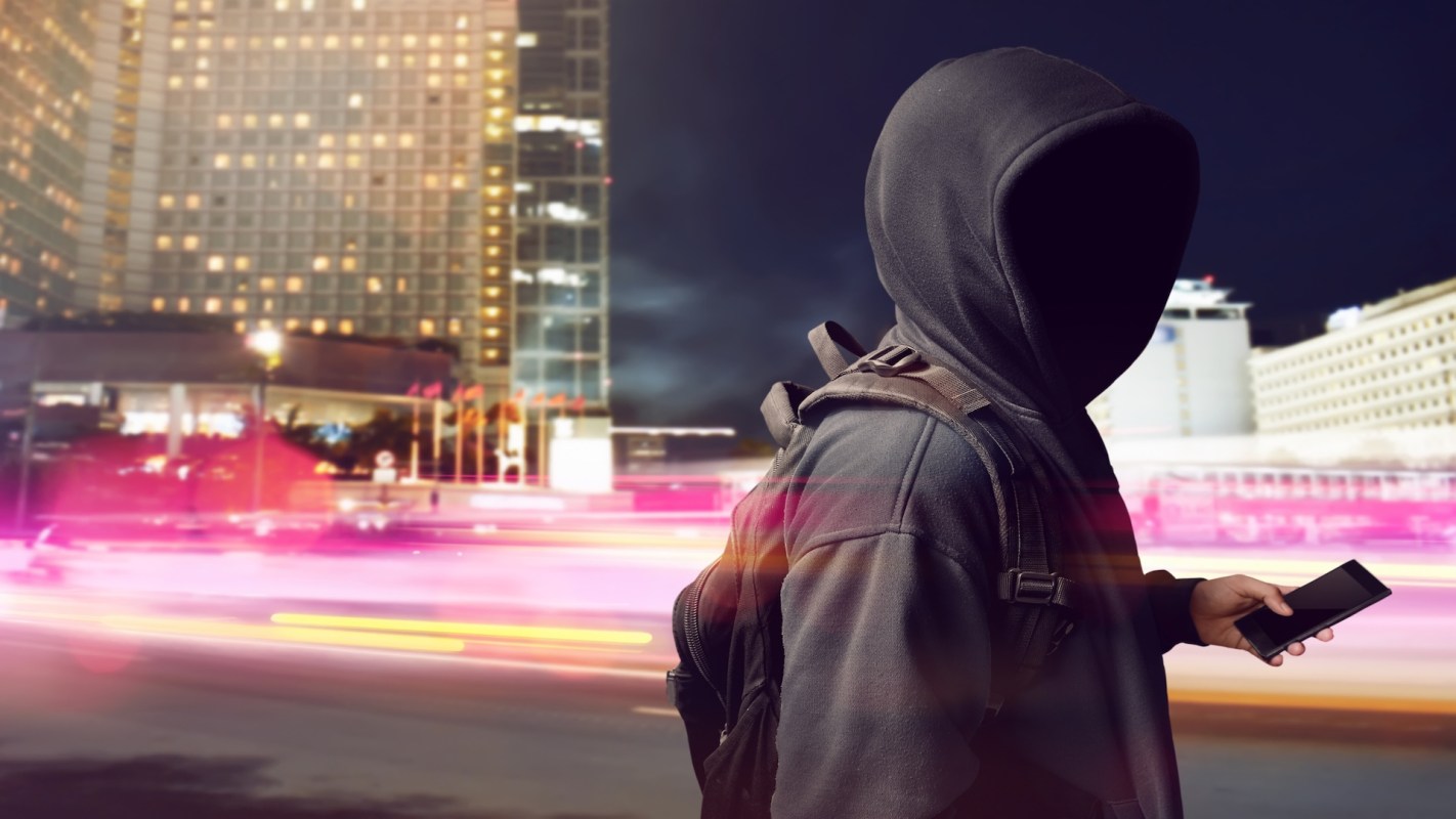 A person in a hoodie stands on a city street at night, holding a smartphone with blurred lights in the background.