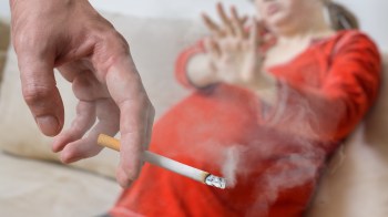 A hand holds a cigarette while a pregnant woman in a red top appears to push away from the smoke.