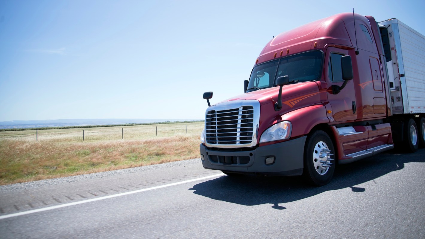 A red semitruck driving on a highway with fields in the background.