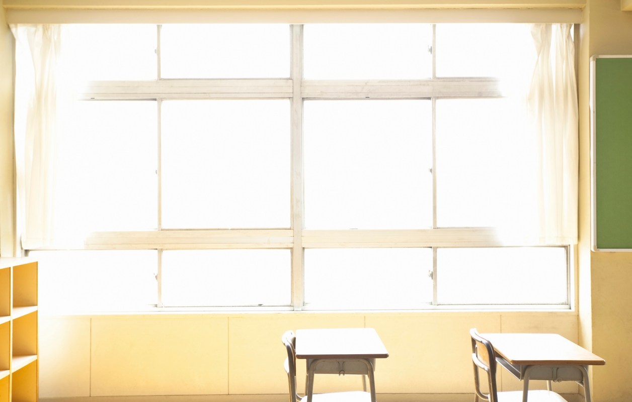 Two empty desks near a large window in a bright classroom with a green chalkboard on the wall.