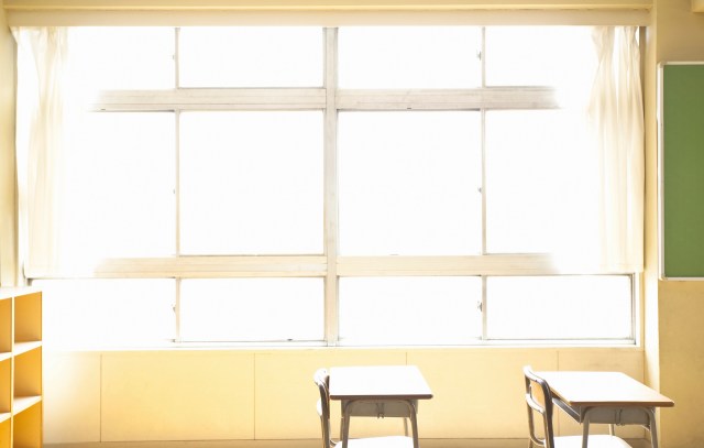 Two empty desks near a large window in a bright classroom with a green chalkboard on the wall.