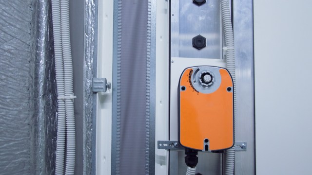 An orange control device mounted on a wall near white tubing and silver insulation material.