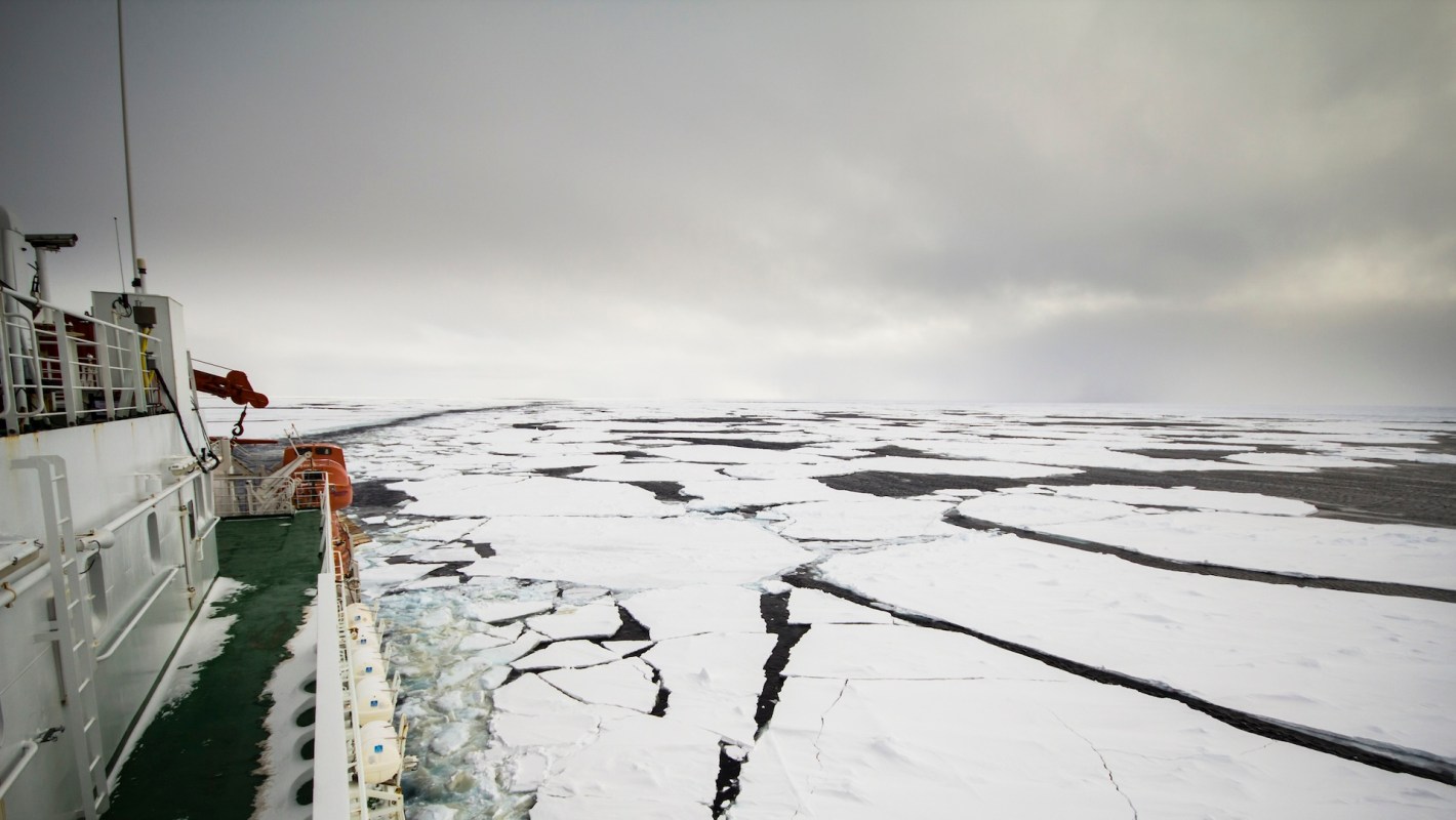 A ship navigates through a frozen sea with large patches of ice and open water under a cloudy sky.