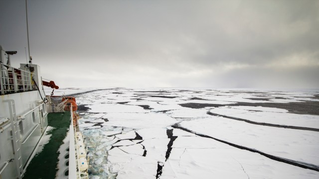 A ship navigates through a frozen sea with large patches of ice and open water under a cloudy sky.