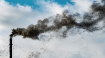 A smokestack emits thick black smoke into a blue sky with white clouds.