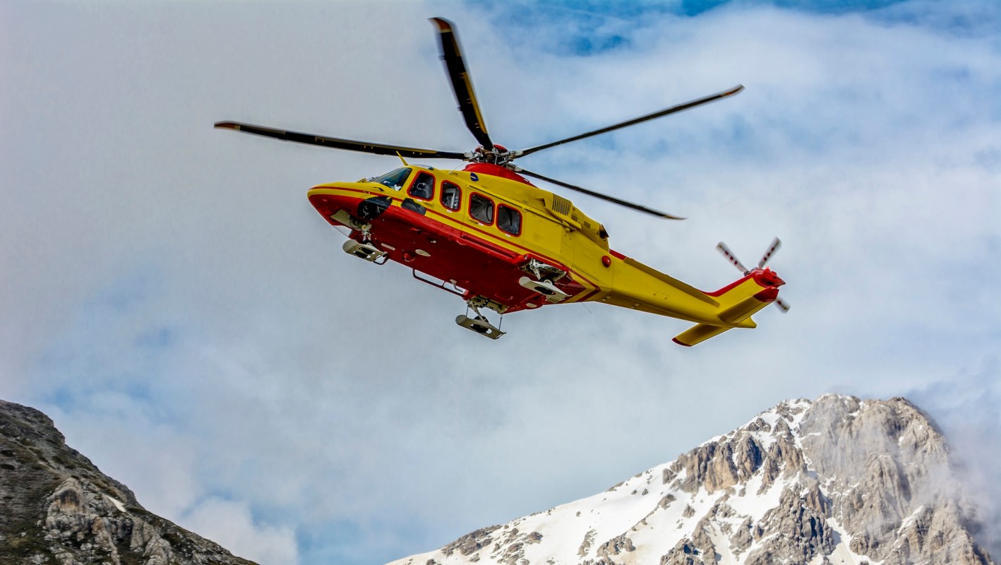 A yellow-and-red rescue helicopter flies over snow-capped mountains under a cloudy sky.