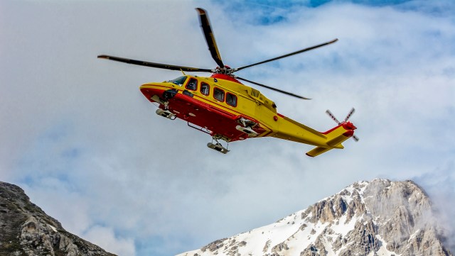 A yellow-and-red rescue helicopter flies over snow-capped mountains under a cloudy sky.