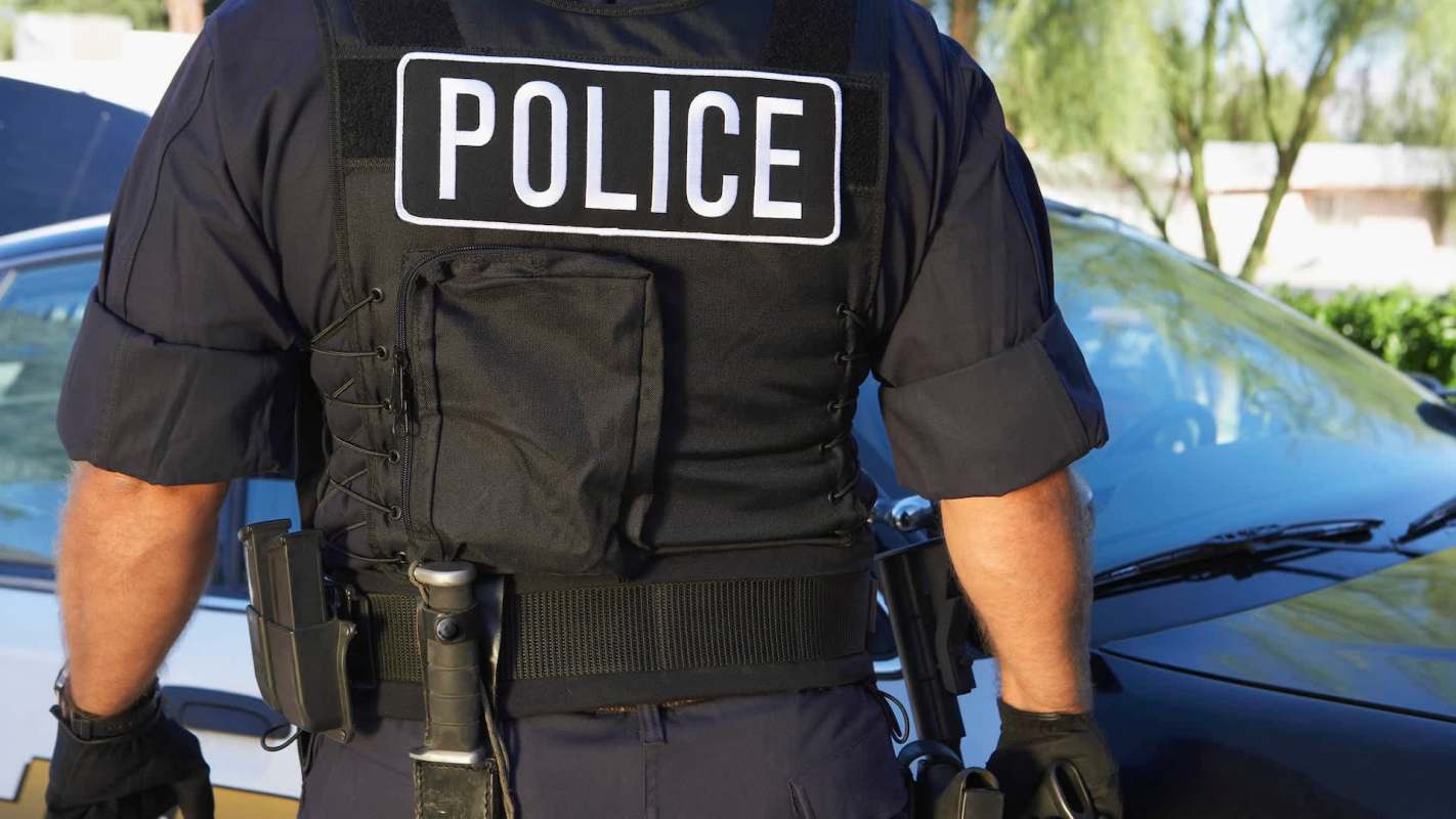 A police officer stands next to a patrol car wearing a tactical vest.
