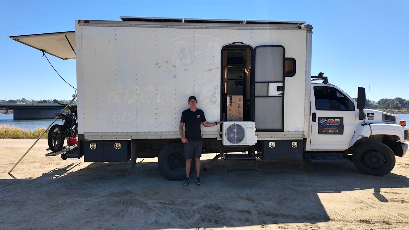 A man stands next to a truck with an air conditioning unit and an open door revealing boxes inside.
