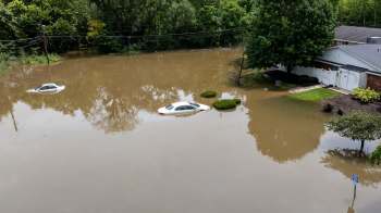 Two cars partially submerged in floodwater near a house surrounded by trees and shrubs.