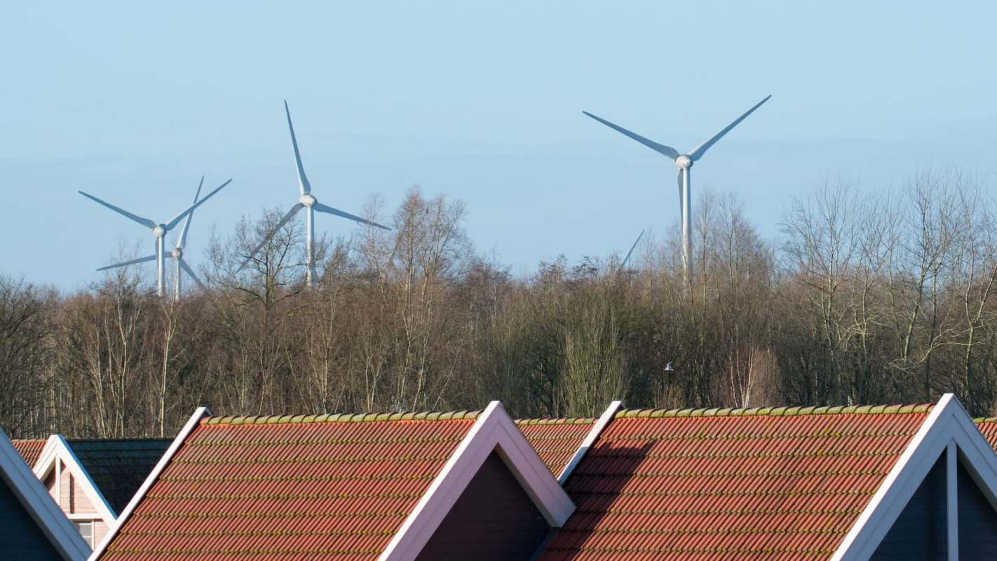 Wind turbines are visible above the rooftops of residential houses and trees.