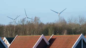 Wind turbines are visible above the rooftops of residential houses and trees.