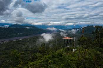 A lookout tower overlooks a dense forest and mountain range under a cloudy sky.