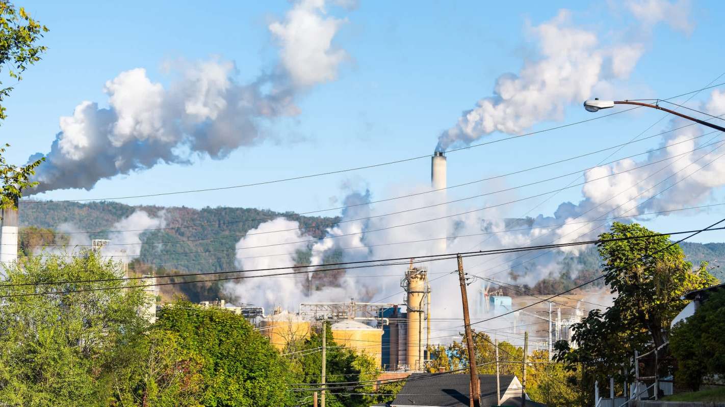 Industrial smokestacks release clouds of smoke against a blue sky and distant hills, with power lines in the foreground.