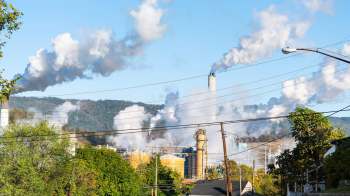 Industrial smokestacks release clouds of smoke against a blue sky and distant hills, with power lines in the foreground.