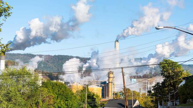 Industrial smokestacks release clouds of smoke against a blue sky and distant hills, with power lines in the foreground.