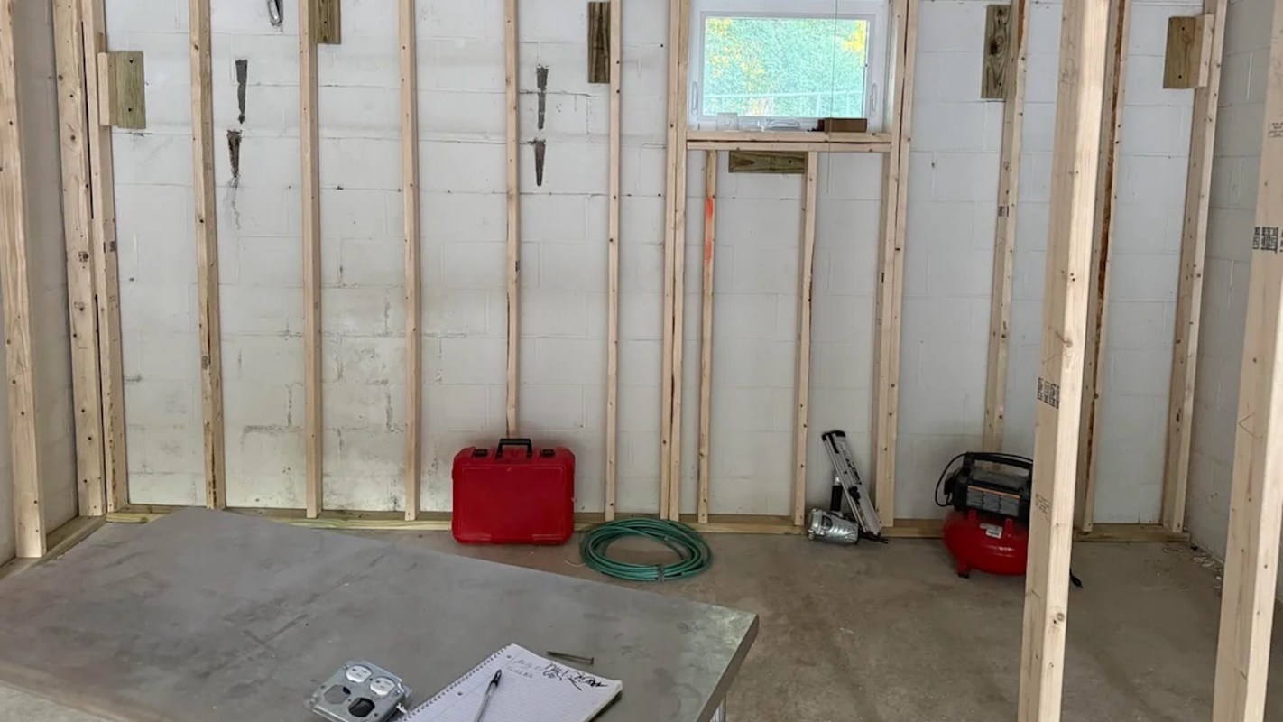 An unfinished room with wooden framing, a small window, tools, a red toolbox, and a coiled green hose.