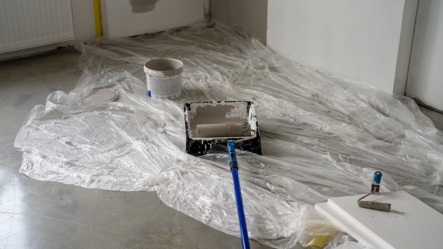 A paint roller and tray sit on a sheet of plastic in a room undergoing painting.