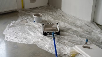A paint roller and tray sit on a sheet of plastic in a room undergoing painting.
