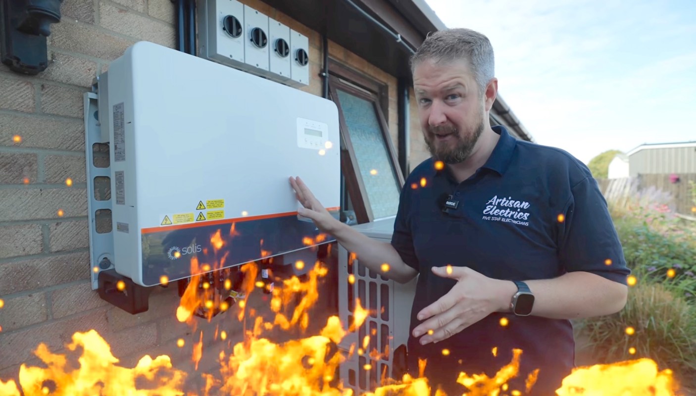 A man points to a solar inverter mounted on a brick wall with a graphic of orange flames in the foreground.