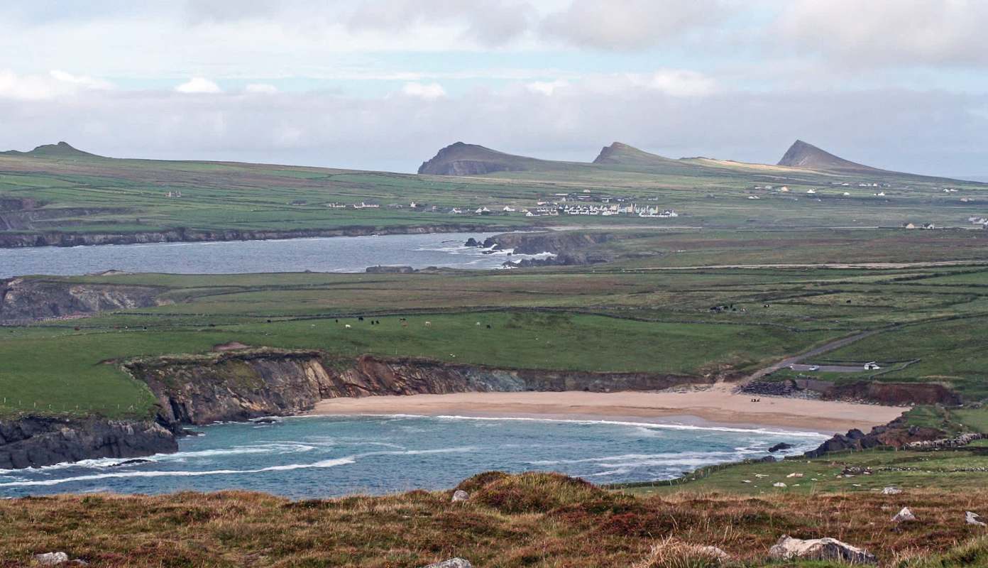 A coastal landscape with a sandy beach, rolling green hills, and distant mountains under a cloudy sky.