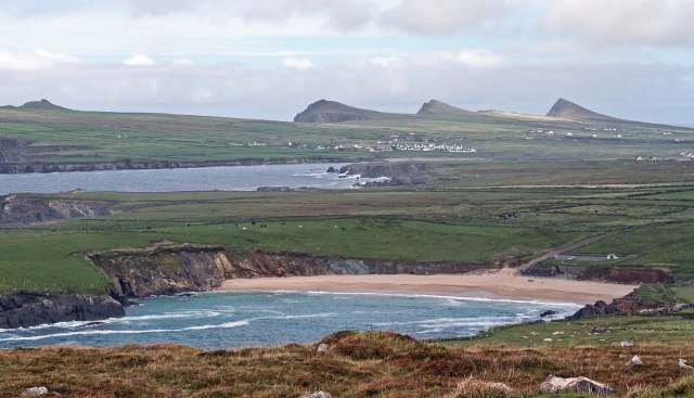 A coastal landscape with a sandy beach, rolling green hills, and distant mountains under a cloudy sky.