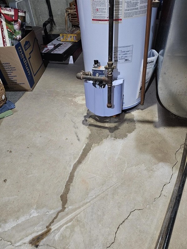 A water heater on a concrete floor with visible water stains and drainage pipes.