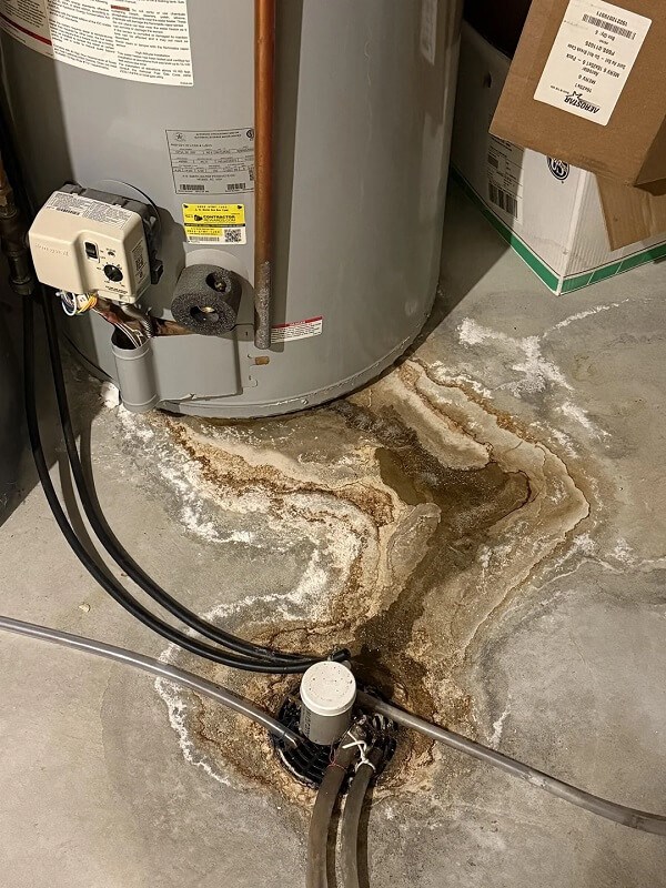 Water heater with a stained floor and drainage pipe, showing signs of water damage and mineral deposits.