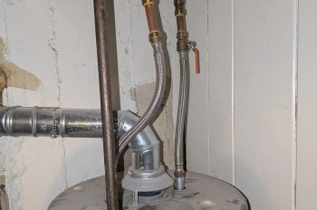 Pipes and hoses connect to a water heater in a basement, with a visible shut-off valve and wall.