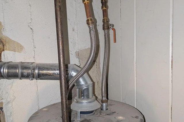 Pipes and hoses connect to a water heater in a basement, with a visible shut-off valve and wall.