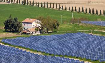 A house near a field of solar panels with trees and a path in the background.