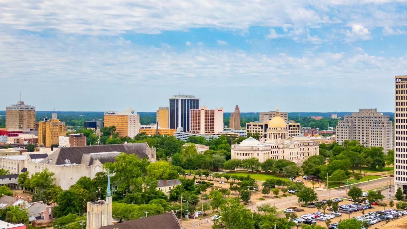 A skyline of Jackson, Mississippi, featuring high-rise buildings, green parks, and a prominent dome-shaped structure.