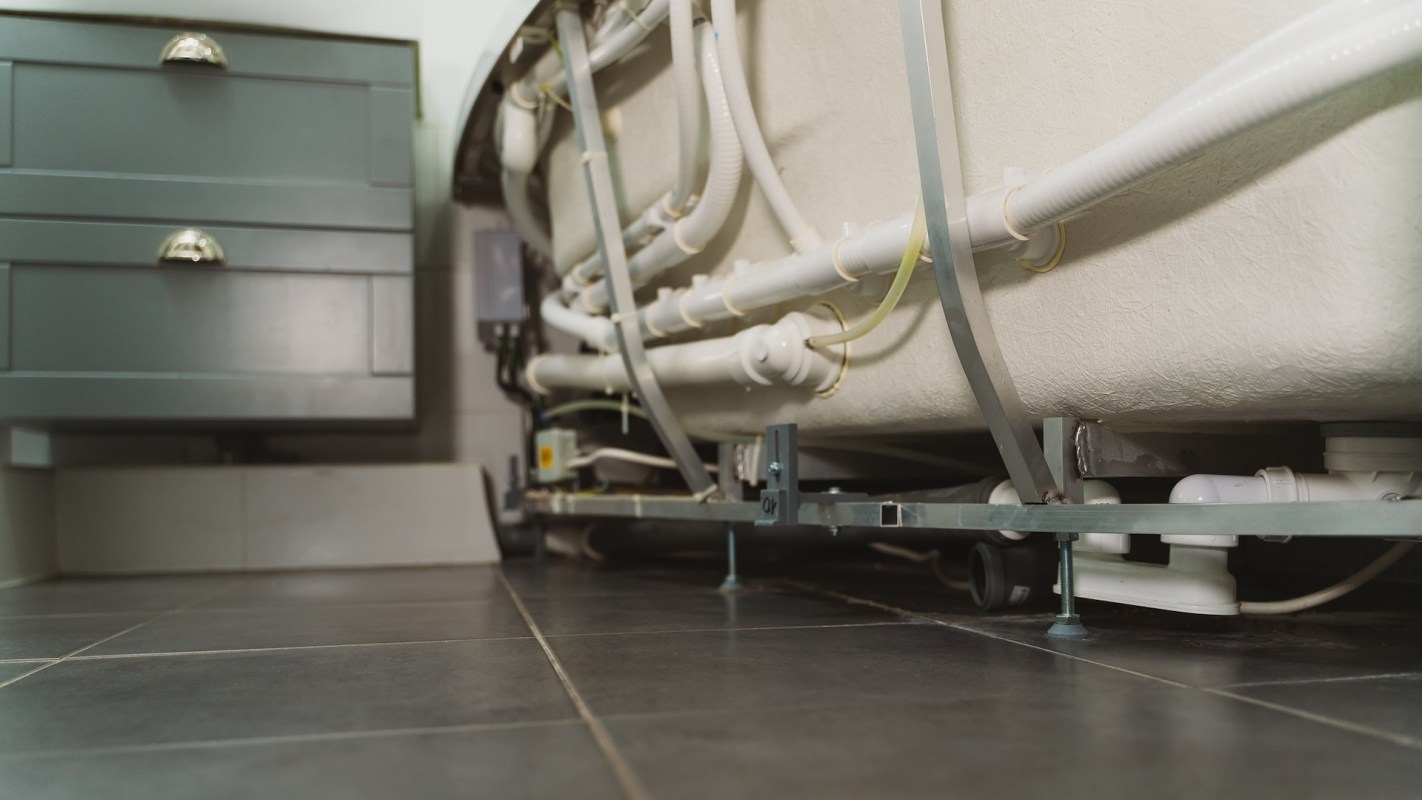Pipes and tubing are visible beneath a bed frame, alongside a gray cabinet and tiled floor.