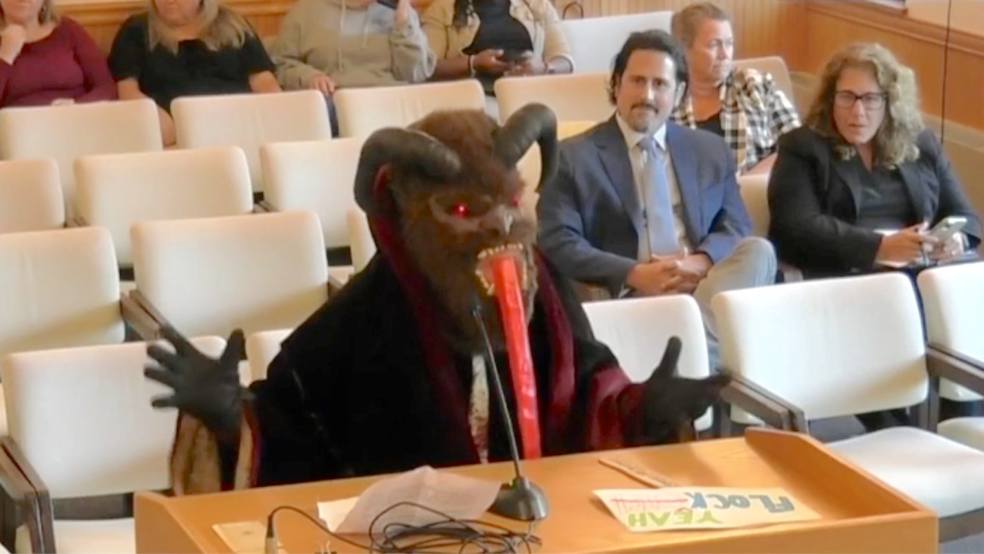 A person in a creature disguise with horns and red eyes gestures while seated at a table in a meeting room.