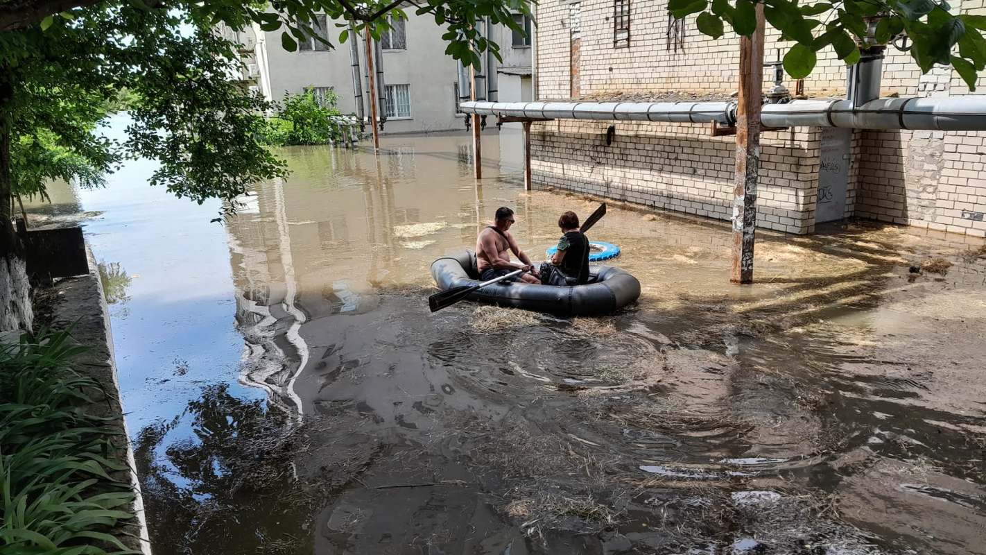 Two people in a raft navigate through floodwaters near a building with overgrown vegetation.