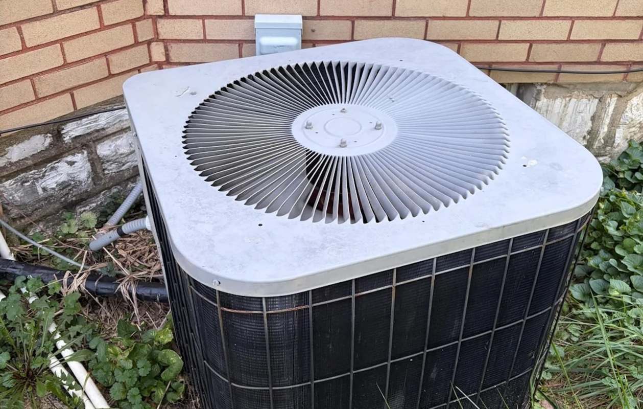 An outdoor air conditioning unit beside a brick wall with greenery around it.