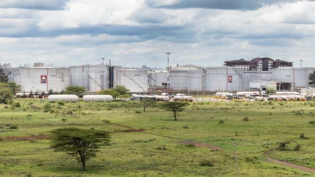 An oil storage facility with multiple large tanks and trucks, set in a grassy area under a cloudy sky.