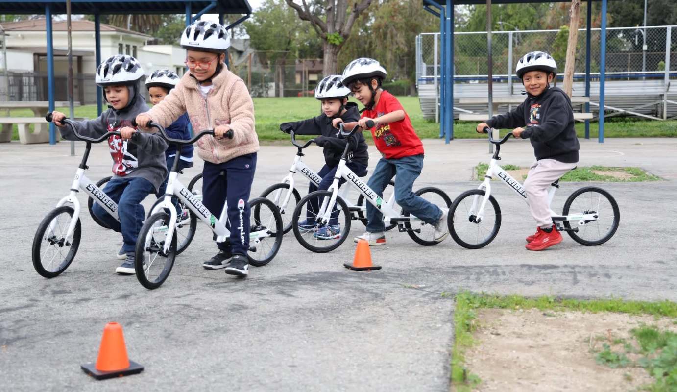 A group of children riding balance bikes in a playground with cones on the ground.
