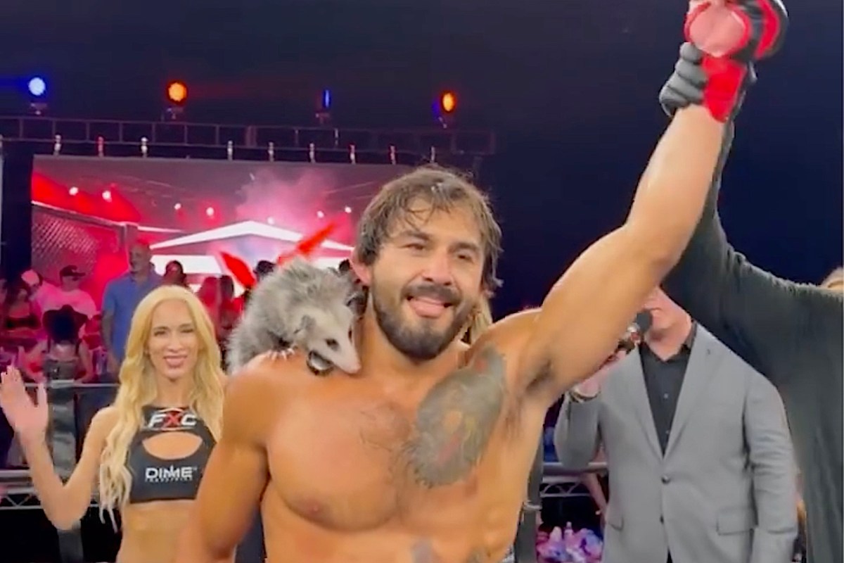 A man in a wrestling ring holds a championship belt with an opossum on his shoulder, while a woman waves in the background.