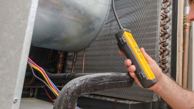 A person using a testing device on an HVAC unit with exposed wiring and copper tubing.