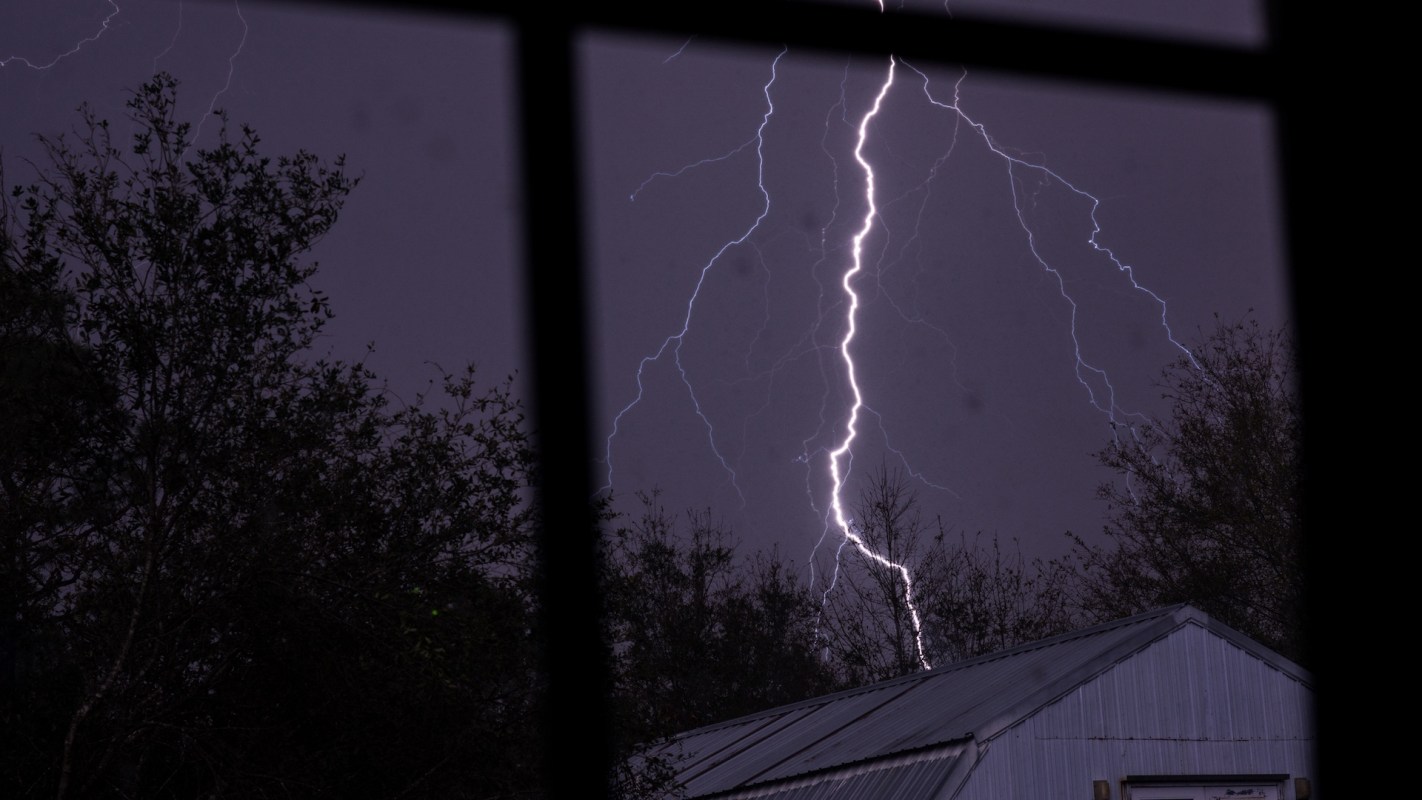 A lightning strike illuminates a dark sky above trees and a building.