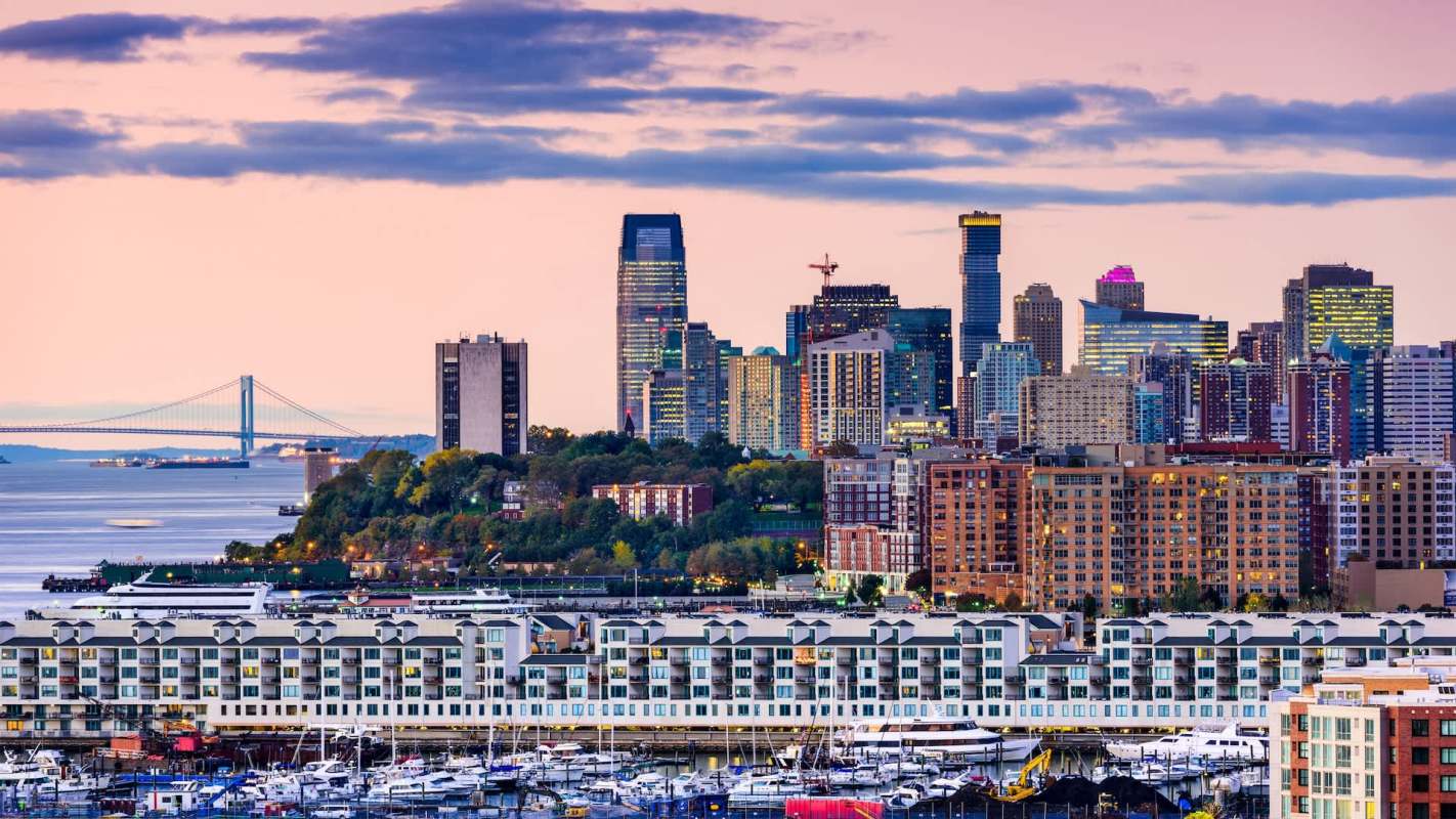 A waterfront city skyline with buildings, a bridge, and boats moored at a marina.