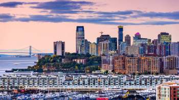 A waterfront city skyline with buildings, a bridge, and boats moored at a marina.