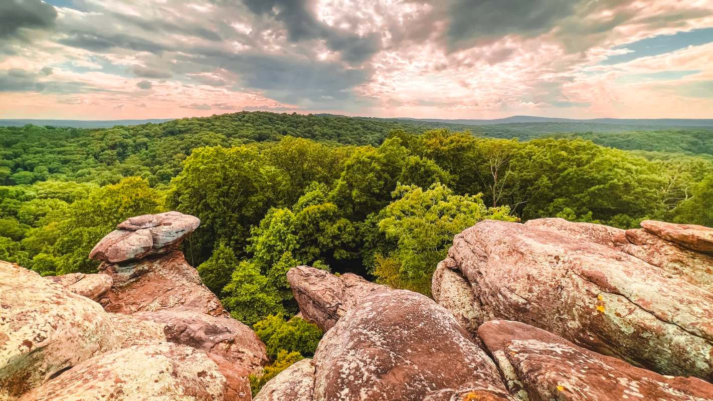 Rocks overlook a dense, green forest under a cloudy sky with light peeking through.