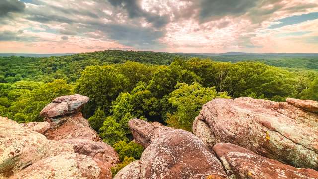 Rocks overlook a dense, green forest under a cloudy sky with light peeking through.