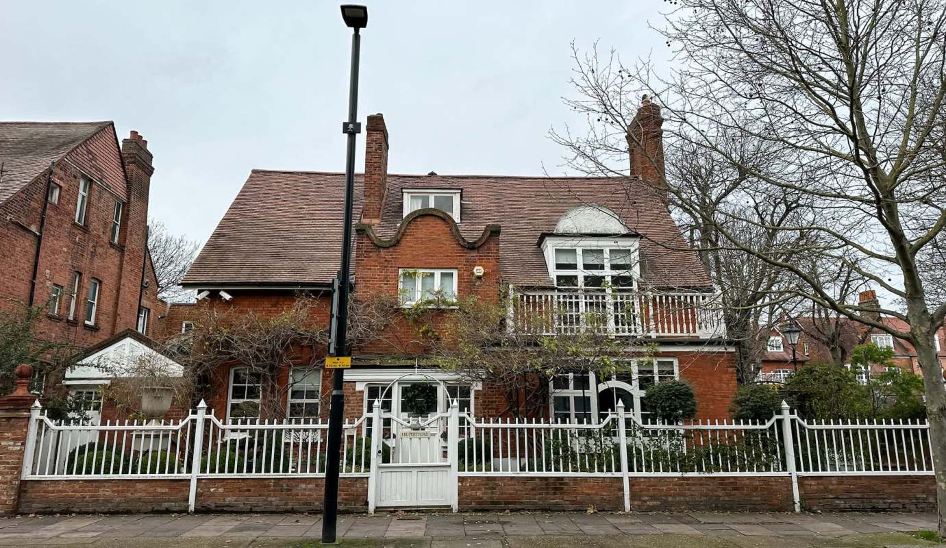 A red brick house with a distinctive rooftop design and a white picket fence, surrounded by trees.