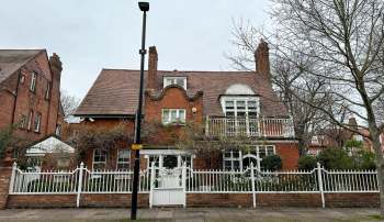 A red brick house with a distinctive rooftop design and a white picket fence, surrounded by trees.