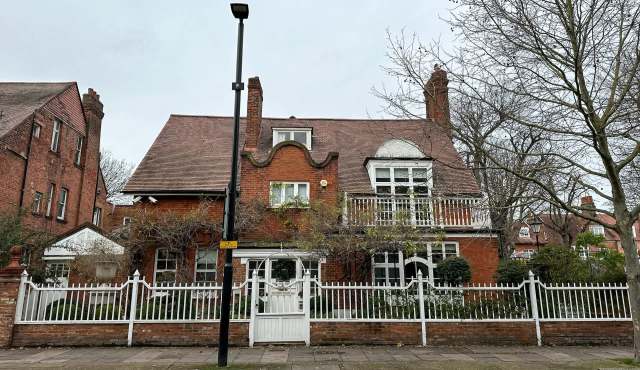 A red brick house with a distinctive rooftop design and a white picket fence, surrounded by trees.