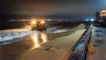 A beach vehicle working at night to manage sand along the shoreline with city lights in the background.