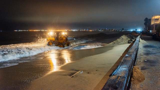 A beach vehicle working at night to manage sand along the shoreline with city lights in the background.